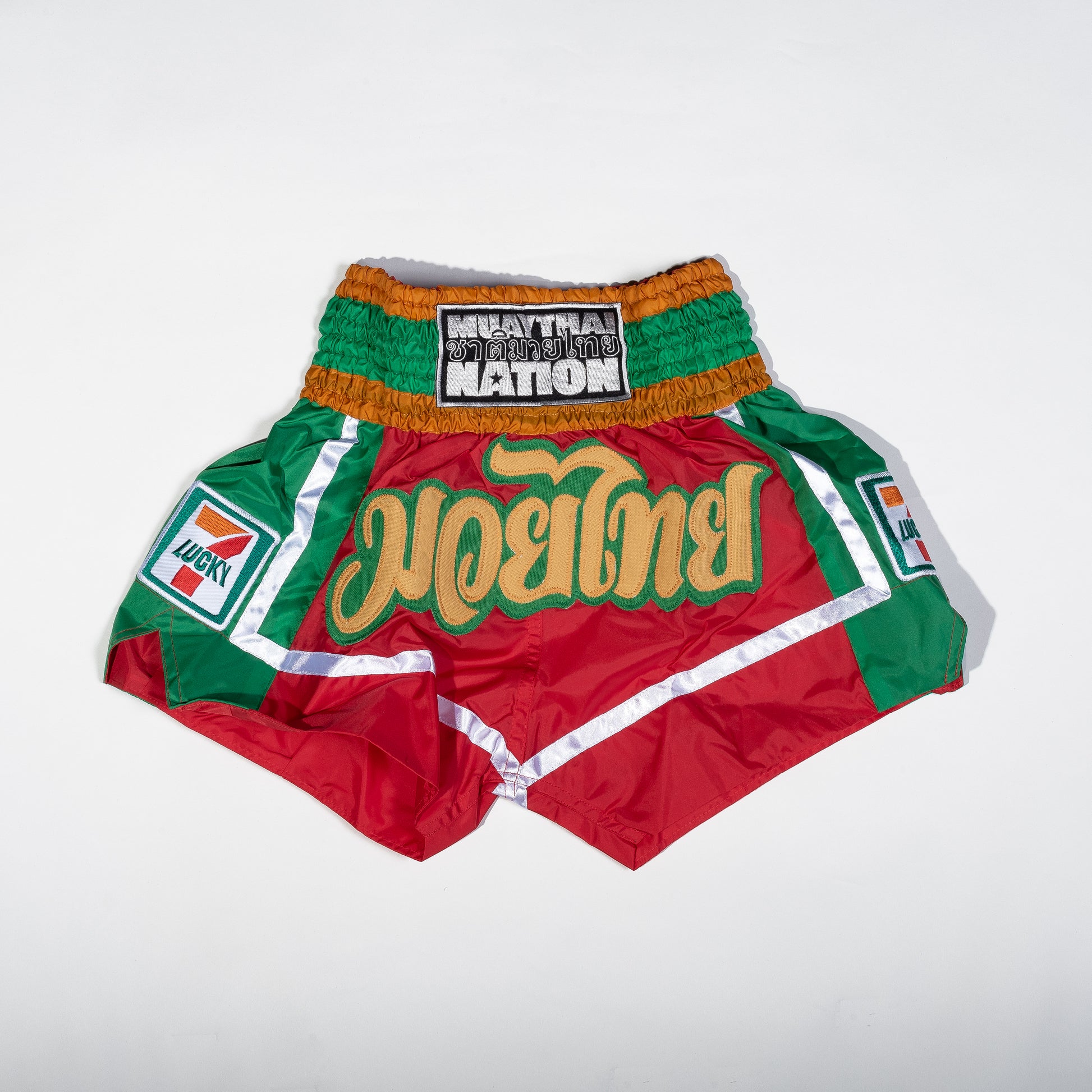 Muay Thai Nation limited edition Lucky 7 red and green satin shorts featuring gold Thai script lettering and a retro convenience store patch design